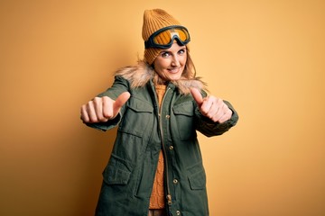 Middle age beautiful blonde skier woman wearing snow sportwear and ski goggles approving doing positive gesture with hand, thumbs up smiling and happy for success. Winner gesture.