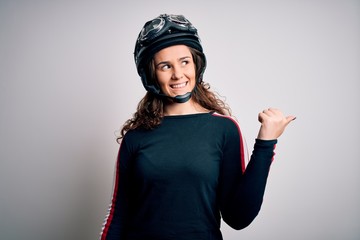 Beautiful motorcyclist woman with curly hair wearing moto helmet over white background smiling with happy face looking and pointing to the side with thumb up.