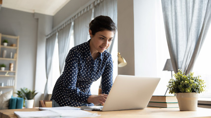 Smiling Indian girl standing at desk, working on laptop, looking at screen, browsing, searching...
