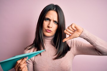 Young woman with blue eyes holding paper airplane standing over isolated pink background with angry face, negative sign showing dislike with thumbs down, rejection concept
