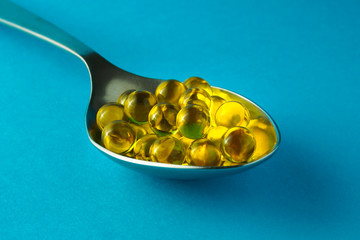 Heap of round fish oil capsules in metallic spoon on blue background, selective focus, close-up.