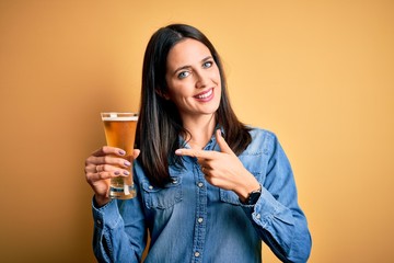 Young woman with blue eyes drinking glass of beer standing over isolated yellow background very...
