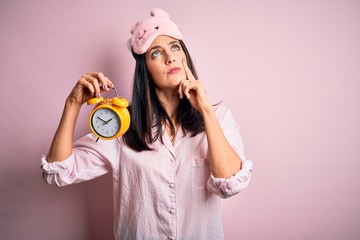 Young brunette woman with blue eyes wearing pajama and eye mask holding alarm clock serious face thinking about question, very confused idea