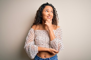 Young beautiful woman with curly hair wearing casual t-shirt standing over white background with hand on chin thinking about question, pensive expression. Smiling with thoughtful face. Doubt concept.