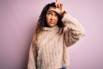 Young beautiful woman with curly hair wearing casual sweater standing over pink background making fun of people with fingers on forehead doing loser gesture mocking and insulting.