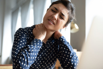 Unhappy Indian girl massaging stiff neck muscles close up, exhausted tired woman sitting at desk...