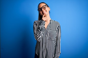 Beautiful blonde woman with blue eyes wearing striped shirt and glasses over blue background looking confident at the camera with smile with crossed arms and hand raised on chin. Thinking positive.