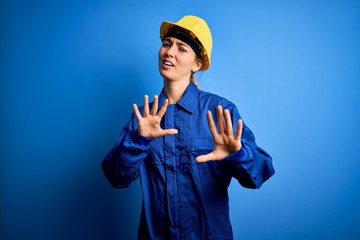 Young beautiful worker woman with blue eyes wearing security helmet and uniform afraid and terrified with fear expression stop gesture with hands, shouting in shock. Panic concept.