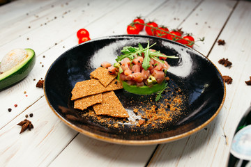 Crispy wrenches and a salad of fresh salmon, arugula and avocado lie on a black plate. Near a wooden background are a bottle, avocado and cherry tomatoes.