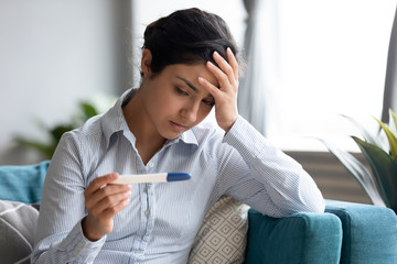 Unhappy Indian woman looking at pregnancy test close up, upset by result, stressed thoughtful young female touching forehead, sitting on couch, unwanted pregnancy or health problem, infertility