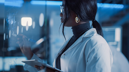In the Research Laboratory Smart and Beautiful African American Female Scientist Wearing White Coat and Protective Glasses Writes Formula on Glass Whiteboard, References Her Tablet Computer