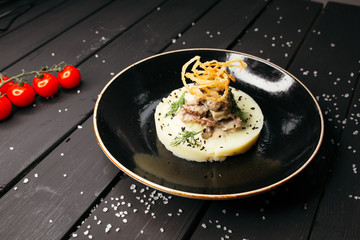Meat on a plate of tender mashed potatoes on a black glossy plate. The black table is sprinkled with salt and decorated with cherry tomatoes.