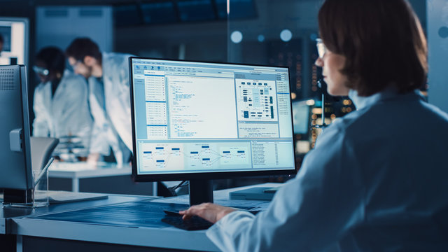 Over The Shoulder Shot: Female IT Scientist Uses Computer Showing System Monitoring And Controlling Program. In The Background Technology Development Laboratory With Scientists, Engineers Working