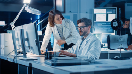 Machine Engine Development Engineer Working on Computer at His Desk, Talks with Project Manager, Shows Prototype Component. Team of Professionals Working in the Modern Industrial Design Laboratory