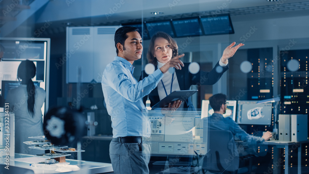 Poster In Technology Research Facility: Female Project Manager Talks With Chief Engineer, they Consult Tablet Computer. Team of Industrial Engineers, Developers Work on Engine Design Using Computers