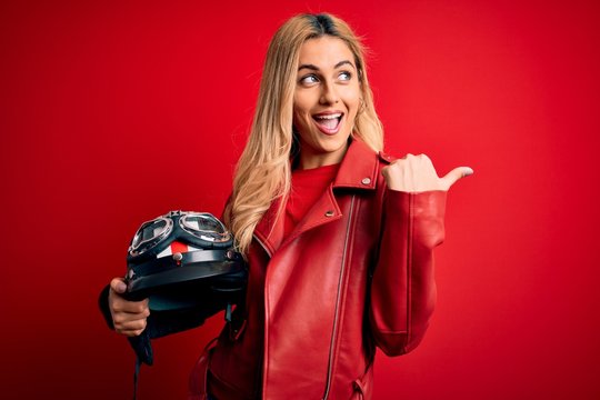 Young beautiful blonde motorcyclist woman holding moto helmet over isolated red background pointing and showing with thumb up to the side with happy face smiling