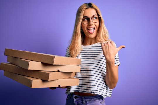 Young beautiful blonde woman holding boxes of italian pizza over isolated purple background pointing and showing with thumb up to the side with happy face smiling