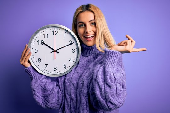 Young beautiful blonde woman doing countdown holding big clock over purple background very happy pointing with hand and finger to the side
