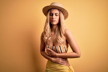 Young beautiful blonde woman on vacation wearing bikini and hat over yellow background with hand on stomach because indigestion, painful illness feeling unwell. Ache concept.