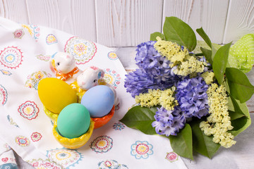 Easter composition with colorful eggs in a ceramic stand-rabbit and spring bouquet with purple and yellow flowers on a white wooden table