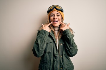 Young brunette skier woman wearing snow clothes and ski goggles over white background Smiling with open mouth, fingers pointing and forcing cheerful smile