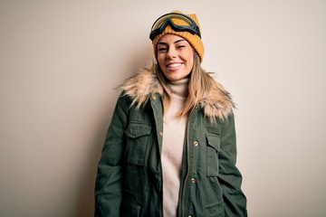 Young brunette skier woman wearing snow clothes and ski goggles over white background winking looking at the camera with sexy expression, cheerful and happy face.