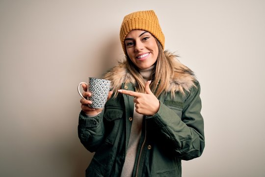 Young beautiful brunette woman wearing snow clothes drinking mug of coffee very happy pointing with hand and finger