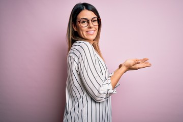 Young beautiful woman wearing casual striped t-shirt and glasses over pink background pointing aside with hands open palms showing copy space, presenting advertisement smiling excited happy