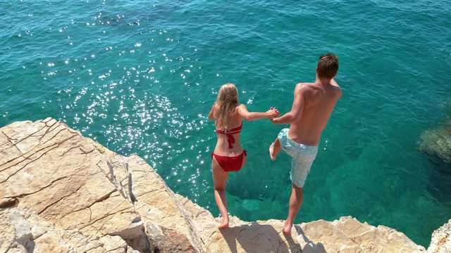 SLOW MOTION: Joyful tourist couple decides to jump off a rocky cliff and dive into sea. Unrecognizable man holds his girlfriend's hand while diving into the deep blue sea on a sunny summer day.