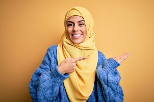 Young beautiful brunette muslim woman wearing arab hijab over isolated yellow background amazed and smiling to the camera while presenting with hand and pointing with finger.