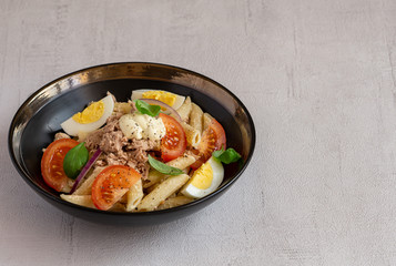 Tuna salad with pasta, tomatoes, and eggs in black bowl isolated on grey background. Healthy, vegetarian dinner or lunch. Italian homemade recipe. Copy space. 