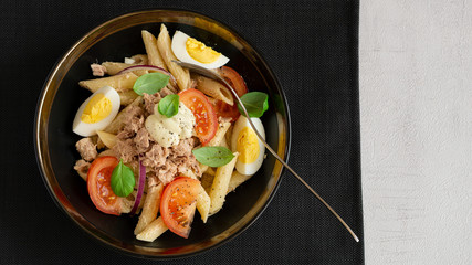 Tuna, pasta, tomato, egg salad in a bowl. Basil leaves. Black mat, fork. Grey background. Top view, copy space. Healthy, vegetarian restaurant dinner, lunch. Recipe, homemade. Italian, mediterranean.