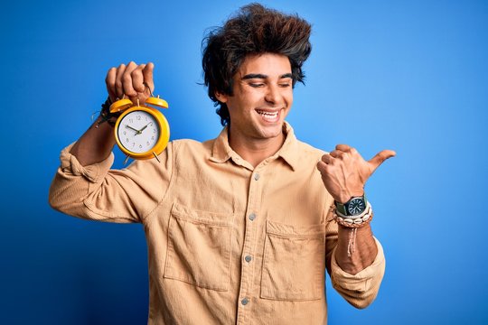 Young handsome man holding alarm clock standing over isolated blue background pointing and showing with thumb up to the side with happy face smiling