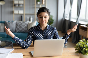 Calm Indian woman with closed eyes meditating at workplace, peaceful mindful young female sitting at desk in modern living room, practicing yoga during break, relieving stress, negative emotions