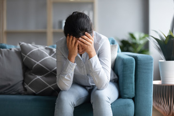 Unhappy depressed Indian woman holding head in hands, sitting alone on couch at home, stressed...