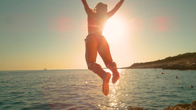 SLOW MOTION, LENS FLARE: Energetic girl jumps off a cliff and kicks legs back while diving into the ocean on a sunny summer evening. Carefree woman does an attractive dive into the refreshing sea.