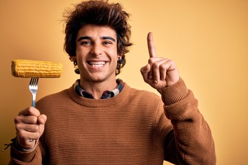 Young handsome man holding fork with cob corn standing over isolated yellow background surprised with an idea or question pointing finger with happy face, number one