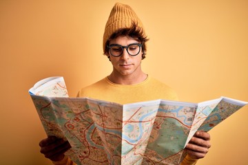 Young tourist man holding map wearing glasses and cap over isolated yellow background with a confident expression on smart face thinking serious