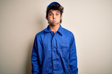 Young mechanic man wearing blue cap and uniform standing over isolated white background puffing cheeks with funny face. Mouth inflated with air, crazy expression.