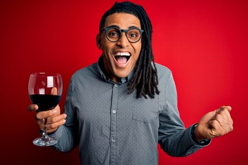 Young african american afro sommelier man with dreadlocks drinking glass of wine screaming proud...