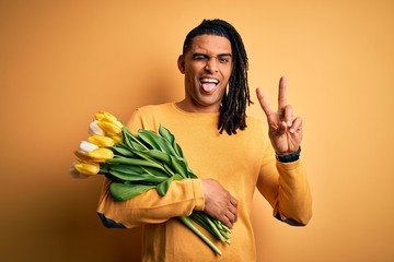 Young african american afro romantic man with dreadlocks holding bouquet of yellow tulips smiling with happy face winking at the camera doing victory sign with fingers. Number two.