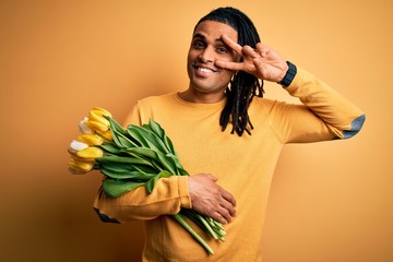 Young african american afro romantic man with dreadlocks holding bouquet of yellow tulips Doing peace symbol with fingers over face, smiling cheerful showing victory