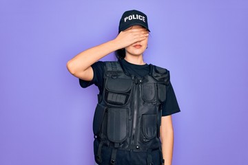 Young police woman wearing security bulletproof vest uniform over purple background covering eyes with hand, looking serious and sad. Sightless, hiding and rejection concept