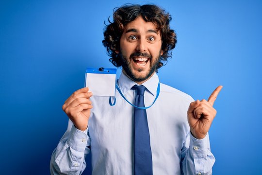 Young Handsome Business Man With Beard Holding Id Card Identification Over Blue Background Very Happy Pointing With Hand And Finger To The Side