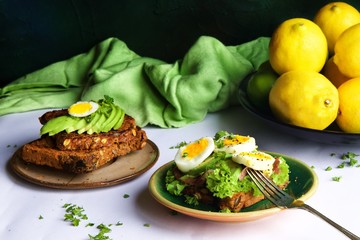 Avocado, capacollo and egg on seed loaf toast.  Lemons and limes in a bowl in the background with a green linen.  Silver fork rests on the side of the plate. Parsley is sprinkled across the scene.