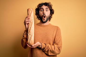Young handsome man with curly hair and beard holding homemade healthy bread scared in shock with a...