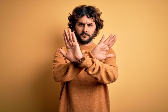 Young handsome man with beard wearing casual sweater standing over yellow background Rejection expression crossing arms and palms doing negative sign, angry face