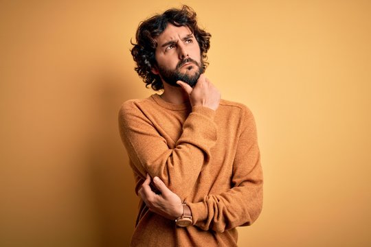 Young handsome man with beard wearing casual sweater standing over yellow background Thinking worried about a question, concerned and nervous with hand on chin