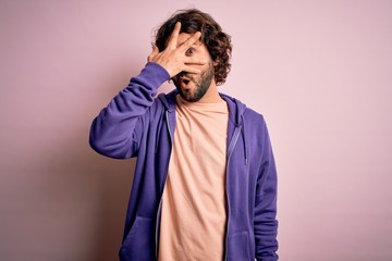 Young handsome sporty man with beard wearing casual sweatshirt over pink background peeking in shock covering face and eyes with hand, looking through fingers with embarrassed expression.