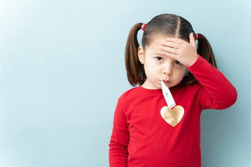 Pretty little girl with ponytails touching her forehead and using a thermometer to check for a fever in a studio